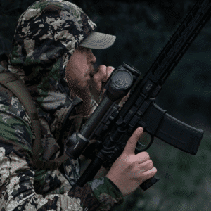 Man in camouflage shown holding an AR15 style rifle with a Pulsar Talion thermal riflescope mounted to it.