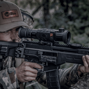 Hunter in camouflage shown looking through the lens of an InfiRay Outdoor RICO Mk1 RH50 V2 shown mounted to a AR15 style rifle.